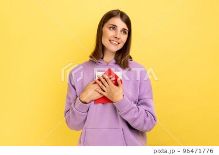 Portrait of beautiful woman standing, embracing envelope and looking away with pleasant smile and enjoying, wearing purple hoodie. Indoor studio shot isolated on yellow background. 96471776