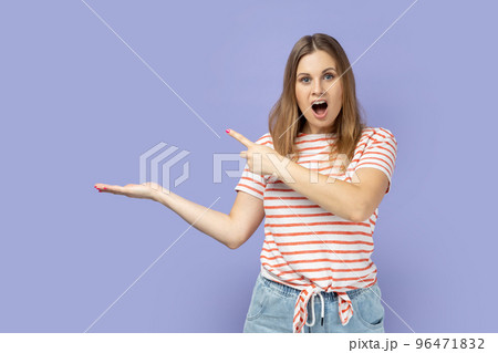 Portrait of surprised astonished blond woman wearing striped T-shirt presenting advertising area on her palm and pointing to copy space. Indoor studio shot isolated on purple background 96471832