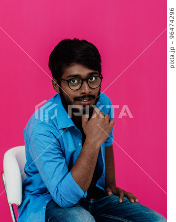A young Indian student in a blue shirt with glasses sitting in front of a pink background A young Indian student in a blue shirt with glasses sitting in front of a pink background 96474296