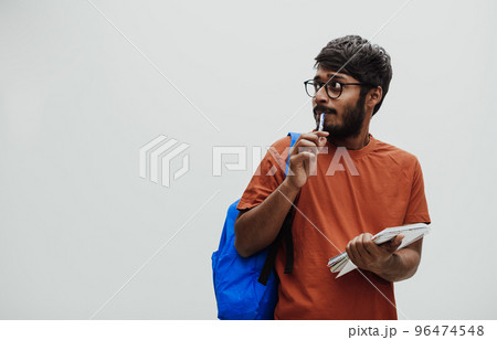 Confused indian student with blue backpack, glasses and notebook posing on gray background. The concept of education and schooling. Time to go back to school Confused indian student with blue backpack, glasses and notebook posing on gray background. The concept of education and schooling. Time to go back to school 96474548