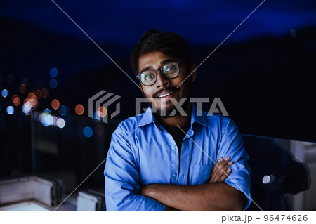An Indian man with glasses and a blue shirt looks around the city at night. In the background of the night street of the city An Indian man with glasses and a blue shirt looks around the city at night. In the background of the night street of the city 96474626