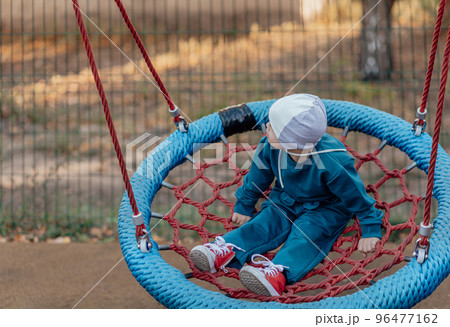 Cute Little boy with down syndrome in a funny hat walks in the playground, swinging on a swing Cute Little boy with down syndrome in a funny hat walks in the playground, swinging on a swing 96477162