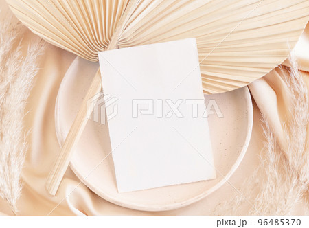 Blank card on plate on dried palm leaf and beige silky fabric close up, greeting or wedding mockup 96485370