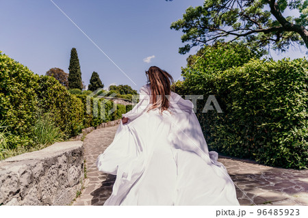 a beautiful woman with long brown hair and long white dress runs along a path along beautiful bushes in the park rear view 96485923