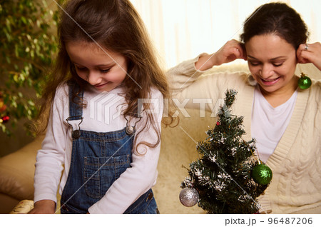 Beautiful Caucasian child, a lovely little girl daughter and her delightful loving mother have fun together, hanging shiny bauble while decorating the Christmas tree at home. Happy New Year concept Beautiful Caucasian child, a lovely little girl daughter and her delightful loving mother have fun together, hanging shiny bauble while decorating the Christmas tree at home. Happy New Year concept 96487206