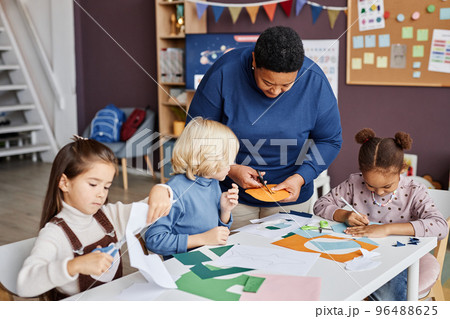 African American mature teaher helping little boy cut holes in paper mask 96488625