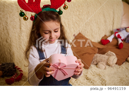 Charming Caucasian little child girl wearing a deer antler hoop, holding a pink heart shaped gift box. Boxing Day. Happy winter holidays. Happy New Year. Merry Christmas. Christmastime 96491159