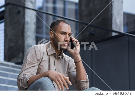 Portrait of a serious and shocked hispanic african american man. He sits gloomily outside on the steps, talks on the phone sadly, looks away. Portrait of a serious and shocked hispanic african american man. He sits gloomily outside on the steps, talks on the phone sadly, looks away. 96492078