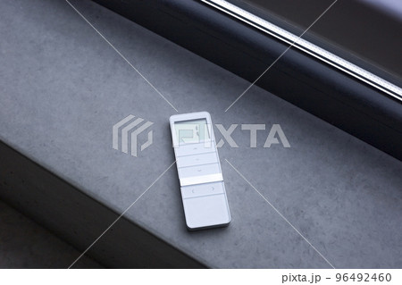 Remote control for window blinds. White remote control lying on a concrete windowsill near the window. Control panel for roller shades and wooden blinds. Remote control for window blinds. White remote control lying on a concrete windowsill near the window. Control panel for roller shades and wooden blinds. 96492460