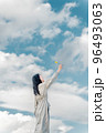 広大な青空のもとで手を伸ばすロングヘアーの女性 96493063