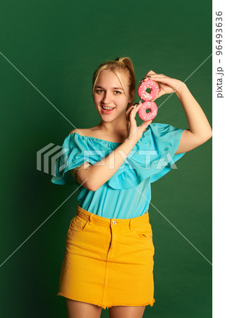 Emotional young beautiful girl, student in summer outfit is goofing around with glazed donuts isolated over green background. Positive emotions, happiness, hobbies and joy Emotional young beautiful girl, student in summer outfit is goofing around with glazed donuts isolated over green background. Positive emotions, happiness, hobbies and joy 96493636