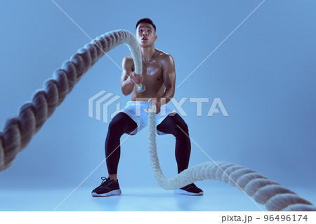 Portrait of young muscular man training, pulling rope isolated over blue background in neon light. Stength and power Portrait of young muscular man training, pulling rope isolated over blue background in neon light. Stength and power 96496174