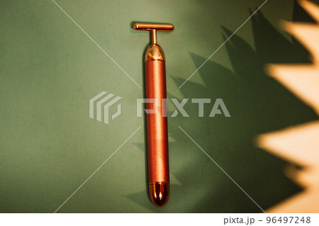 Vibrating golden facial massager, glass jar with oil dropper for massage on emerald background. Female body skin care. Luxury beauty Concept. Remedy for prolonging youthfulness. Pulse face massaging. 96497248