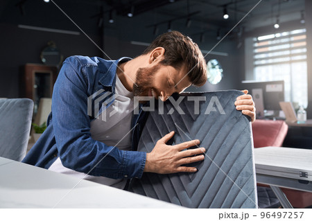 Romantic bearded man in casual outfit touching soft pattern, while leaning to chair indoors. Smiling brunet male with closed eyes hugging grey chair back, while sitting at table. Concept of emotions. 96497257
