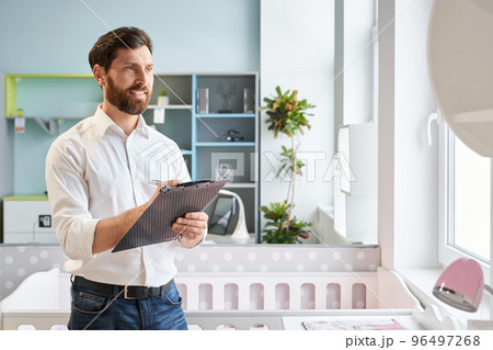 Successful sales manager standing with clip-on tablet in furniture store. Portrait of smiling retailer writing, compare prices, while looking at cupboard in exhibition center. Concept of workplace. Successful sales manager standing with clip-on tablet in furniture store. Portrait of smiling retailer writing, compare prices, while looking at cupboard in exhibition center. Concept of workplace. 96497268