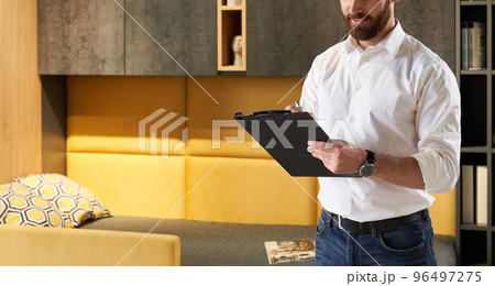 Unrecognizable bearded male retailer making notes, while working in furniture store. Crop view of sales manager writing in tablet, while standing in front of bedroom furniture. Concept of workplace.  96497275