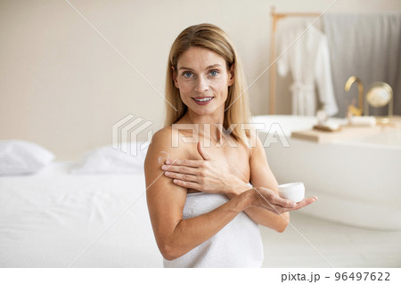 Happy mature lady in bath towel applying cream, putting on moisturizing body butter at home and smiling at camera 96497622