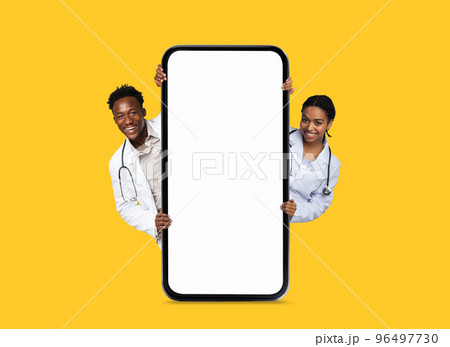 Cheerful black young man and lady doctors in white coats peeking out from big phone with blank screen 96497730
