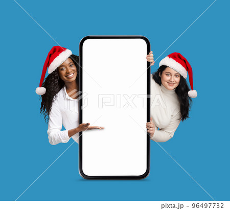 Smiling pretty african american and european millennial women in Christmas hats show hand to big phone 96497732
