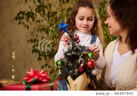 Mother and daughter decorating an eco Christmas tree with beautiful shiny bauble and Xmas toys. Christmastime. New Year's preparations and celebration. Enjoying happy winter holidays in family at home 96497942
