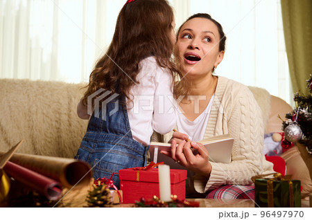 Back view of adorable little girl whispering secrets to her mother as she writes the gift list, while enjoying Christmas and New Year preparations together at home. Happy winter holidays in the family Back view of adorable little girl whispering secrets to her mother as she writes the gift list, while enjoying Christmas and New Year preparations together at home. Happy winter holidays in the family 96497970