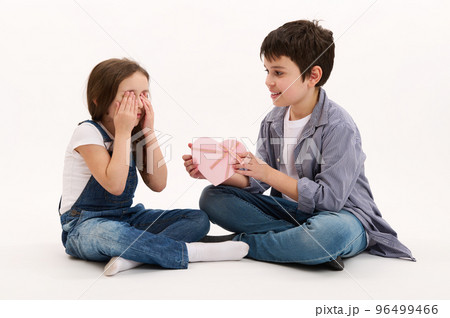 Adorable Hispanic teenage boy, brother gives beautiful heart-shaped pink gift box to his lovely sister, greeting her with St Valentine's Women's Day, isolated on white background. Copy space for text 96499466