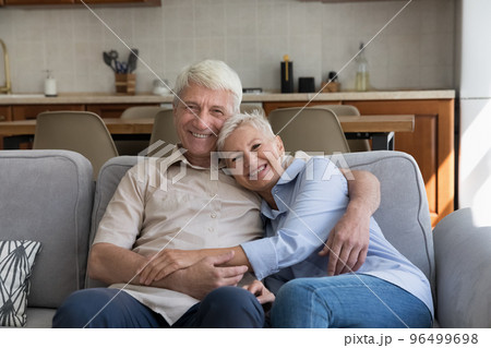 Loving older couple hugging relaxing on sofa looking at camera Loving older couple hugging relaxing on sofa looking at camera 96499698
