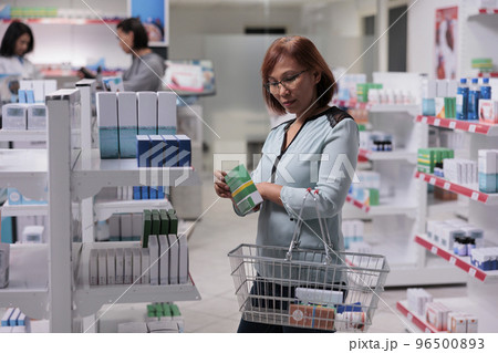 Female customer examining vitamins box on shelves, looking to buy medical supplements and pharmaceutical products. Checking package of healthcare supplies and medicaments in pharmacy shop. Female customer examining vitamins box on shelves, looking to buy medical supplements and pharmaceutical products. Checking package of healthcare supplies and medicaments in pharmacy shop. 96500893