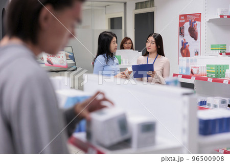 Team of asian women talking about prescription medicine, consultant giving medical advice about pharmaceutical supplements to client. Customer and worker analyzing pills package. 96500908