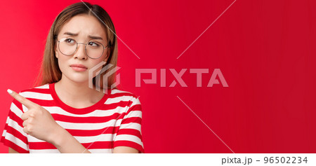 Disappointed doubtful frowning asian girl asking question reassure, looking pointing left displeased, unimpressed with strange place, dislike interiour sulking distressed, stand red background 96502234