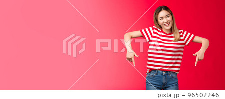 Friendly outgoing attractive blond asian girl pointing down show index fingers bottom advertisement invite guest customers try new product, smiling broadly look cheerful camera, red background 96502246