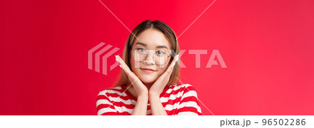 Close-up tender cute asian teenage girl finally got rid pimples skin flaws, touch pure clean skin apply daily skincare products satisfied result, stand pleased red background smiling wear glasses 96502286