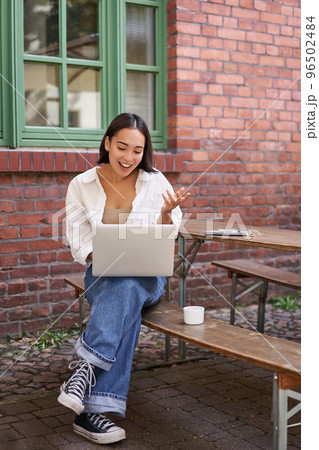 Vertical shot of asian girl with laptop, talking on video call, chatting online, sitting in cafe outdoors 96502484