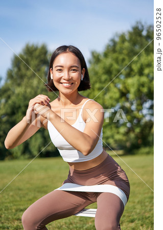 Asian fitness girl doing squats in park, using resistance band, stretching yoga rope for workout training on fresh air Asian fitness girl doing squats in park, using resistance band, stretching yoga rope for workout training on fresh air 96502528