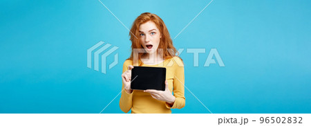 Business Concept - Close up Portrait young beautiful attractive redhair girl smiling showing digital tablet screen on black. Blue Pastel Background. Business Concept - Close up Portrait young beautiful attractive redhair girl smiling showing digital tablet screen on black. Blue Pastel Background. 96502832