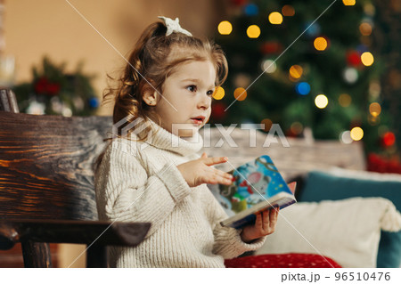 Portrait of a curly-haired little girl in a warm knitted sweater with a book in her hands against the background of a Christmas tree enjoying a cozy winter evening at home. Christmas, New Year 96510476