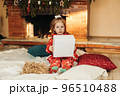 A little girl opens a Christmas present near the Christmas tree. Christmas time, gifts, festive mood. The concept of Christmas and New Year 96510488