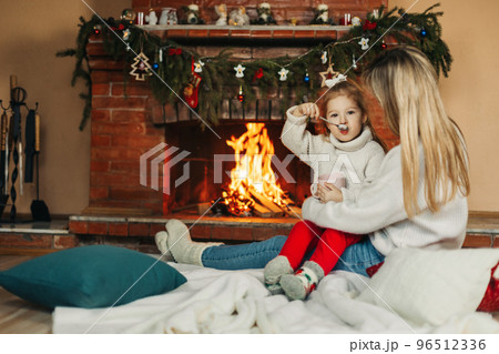 Family mother and child daughter hug and warm up on a winter evening by the fireplace, drink hot cocoa. Christmas Eve or New Year's Eve, Christmas holidays, Christmas time, family evening 96512336