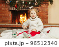 A baby girl drinks hot chocolate by the fireplace at home. A little girl on Christmas Eve or New Year's Eve at home. The concept of Christmas and New Year 96512340