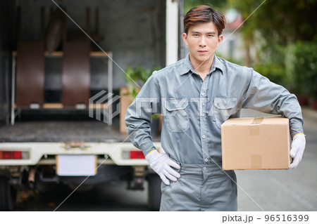 Mover in grey uniform holding cardboard box when standing next to van Mover in grey uniform holding cardboard box when standing next to van 96516399