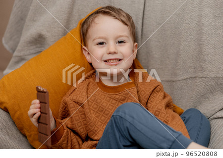 Little adorable boy sitting on the couch at home and eating chocolate bar. Child and sweets, sugar confectionery. Kid enjoy a delicious dessert. Preschool child with casual clothing. Positive emotion. Little adorable boy sitting on the couch at home and eating chocolate bar. Child and sweets, sugar confectionery. Kid enjoy a delicious dessert. Preschool child with casual clothing. Positive emotion. 96520808