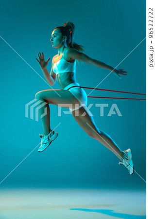 Young sportive girl in white sportswear training with sports expanders isolated over blue studio background in neon light. Run 96522722