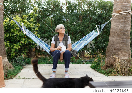 Beautiful senior blonde woman reading book and sitting in hammock in the garden with cat Beautiful senior blonde woman reading book and sitting in hammock in the garden with cat 96527893