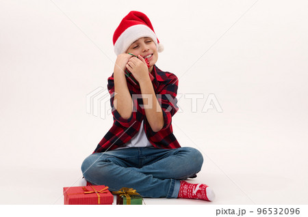 Full length portrait: handsome preteen boy in red plaid shirt and Santa hat, hugging a Christmas gift, sitting on white background with copy space for text. Looking forward to upcoming winter holidays 96532096