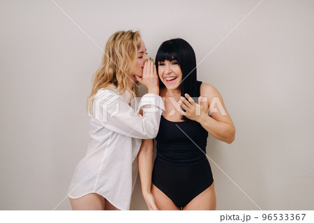 An excited woman in a black swimsuit listens to a friend in a shirt whispering in her ear on a light background 96533367