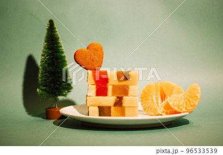 Marzipan marmalade turrone on plate, tangerine on green emerald background. Spanish dessert made from almonds, honey, sugar, eggs. Christmas sweets, heart-shaped cookie. New Year pine tree decor 2023. 96533539