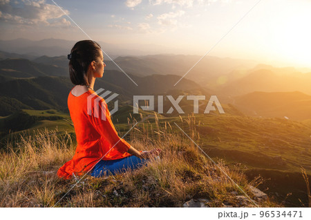 young caucasian woman meditates in the lotus position in the grass in the mountains near the cliff in the setting sun young caucasian woman meditates in the lotus position in the grass in the mountains near the cliff in the setting sun 96534471
