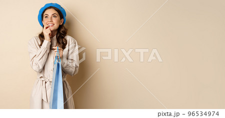 Image of modern beautiful woman posig with shopping gift bag, smiling and looking up thoughtful, thinking, standing over beige background 96534974