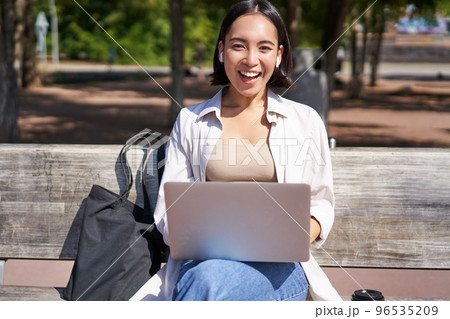 Portrait of asian woman sitting with laptop on bench in park, listening music with wireless headphones, doing homework, working on remote 96535209