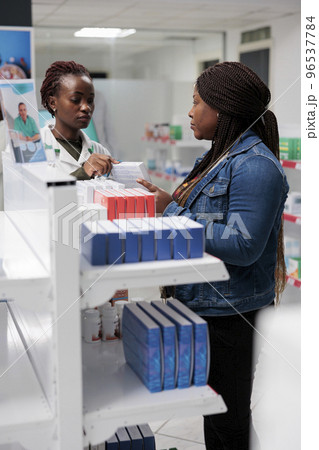 African american pharmacy assistant explaining buyer vitamins instruction in drugstore, woman choosing medications, buying food supplements. Medicaments retail, pharmacist helping client African american pharmacy assistant explaining buyer vitamins instruction in drugstore, woman choosing medications, buying food supplements. Medicaments retail, pharmacist helping client 96537784